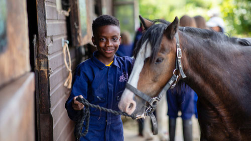 Smiling boy in farm overalls holding horse's bridle.